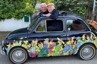 Cartoonist Peter Gaymann und Musik-Kabarettist Josef Brustmann schauen aus einem Auto mit Hühner-Cartoons von Peter Gaymann. 